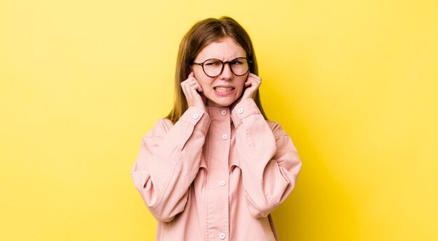 Donna graziosa dalla testa rossa che sembra arrabbiata, stressata e infastidita che copre entrambe le orecchie con un suono assordante o musica ad alto volume