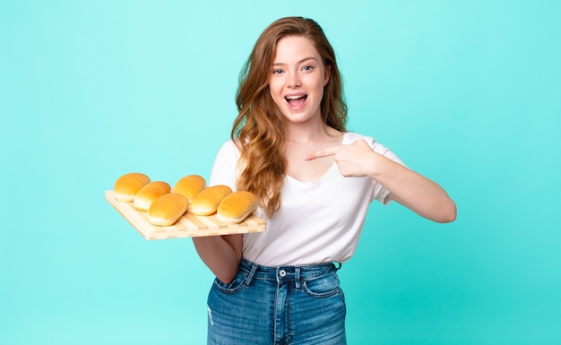 Photo red head pretty woman looking excited and surprised pointing to the side and holding a bread buns troy