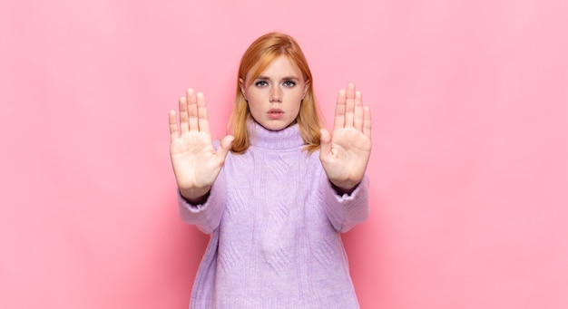 Red head pretty woman looking serious, unhappy, angry and displeased forbidding entry or saying stop with both open palms