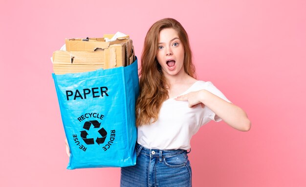 Red head pretty woman looking shocked and surprised with mouth wide open, pointing to self and holding a recycled paper bag