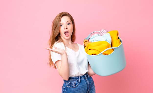 Red head pretty woman looking surprised and shocked, with jaw dropped holding an object and holding a wash basket with clothes