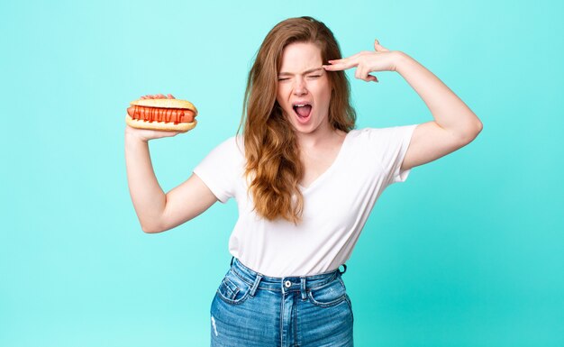 red head pretty woman looking unhappy and stressed, suicide gesture making gun sign and holding a hot dog