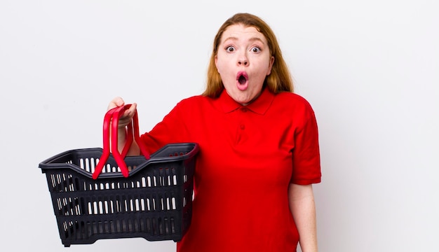 Рыжая красивая женщина выглядит очень шокированной или удивленной концепцией пустой корзины для покупок