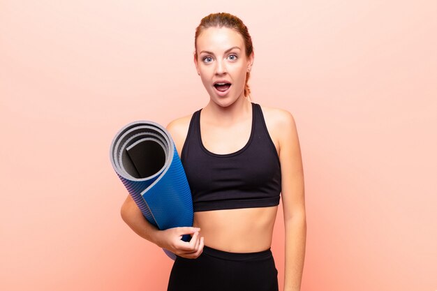 Red head pretty woman looking very shocked or surprised, staring with open mouth saying wow holding a yoga mat.