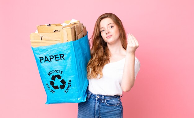 Red head pretty woman making capice or money gesture, telling you to pay and holding a recycled paper bag