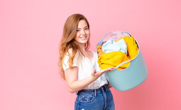 Red head pretty woman smiling cheerfully, feeling happy and showing a concept and holding a wash basket with clothes