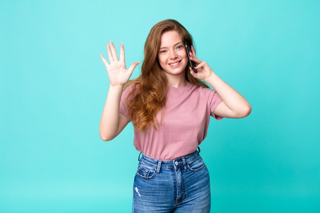 Red head pretty woman smiling and looking friendly, showing number five and using a smartphone