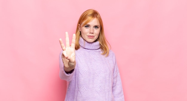 Red head pretty woman smiling and looking friendly, showing number three or third with hand forward, counting down