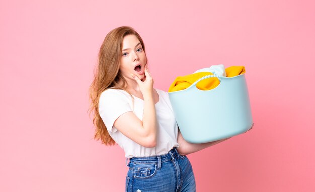 Bella donna dai capelli rossi con la bocca e gli occhi spalancati e la mano sul mento e con in mano un cesto per il bucato con i vestiti