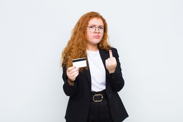 Red head young woman feeling angry, annoyed, rebellious and aggressive, flipping the middle finger, fighting back on white wall