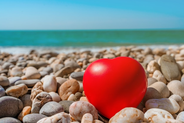 The red heart lies on the pebbles on the beach in front of the azure sea or ocean