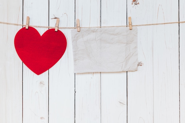 Red heart and old paper blank hanging at clothesline on wood white background with space. Valentine Day.