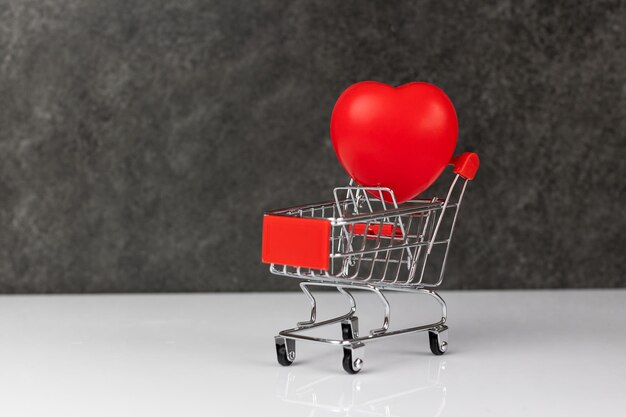 Red heart in shopping cart on a white table on dark grey background