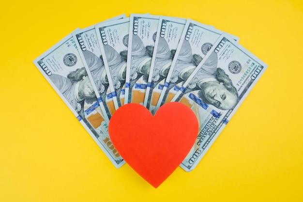 Photo red heart and stack of one hundred dollar bills on a yellow background