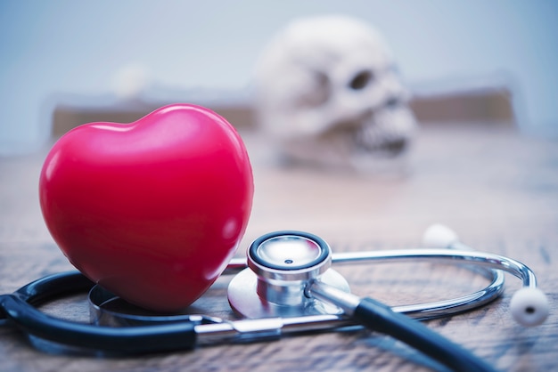  Red heart and stethoscope on wood table with blurred skull on wall background.