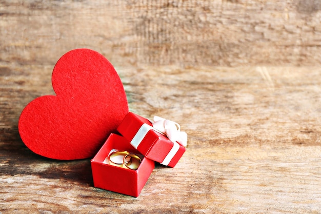 Red heart and wedding rings in a box on wooden background closeup