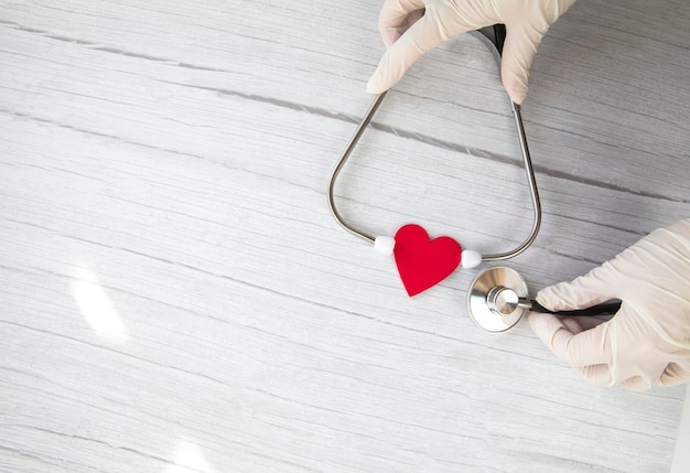 Red heart and white protective medical gloves holds a stethoscope on a white wooden background conce...