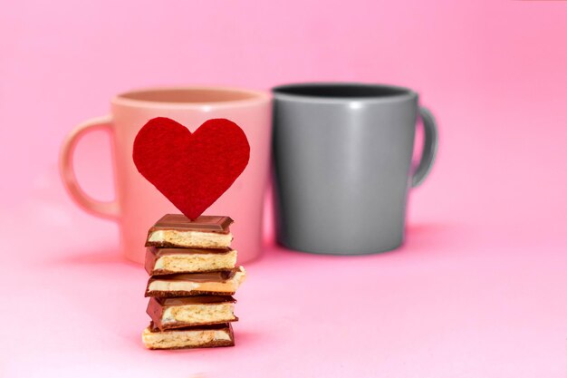 Red heart with chocolate stand on a background of pink and gray cups that stand on a pink background