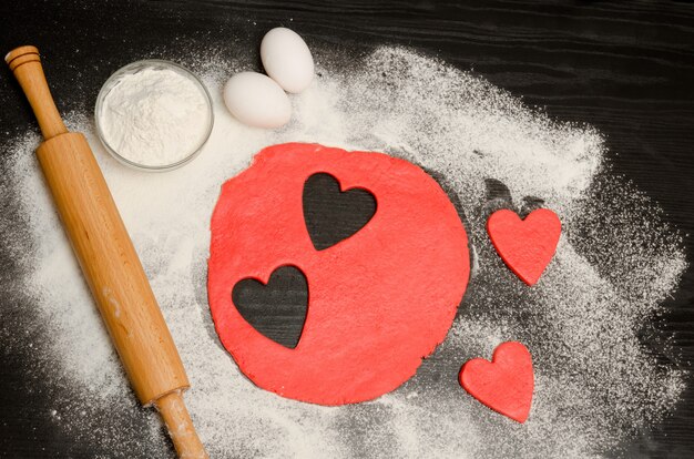 Red hearts cut out dough with rolling pin, eggs and flour on a black table . View from above. copyspace