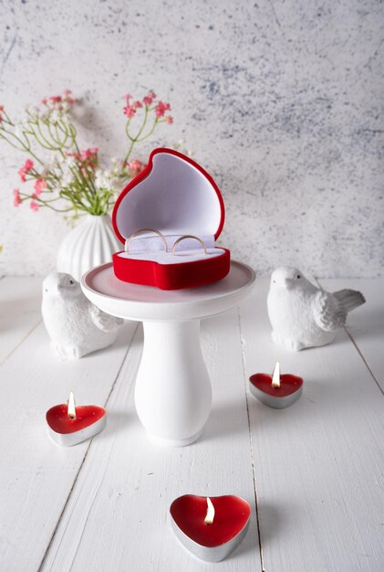 A red heartshaped ring box on a white stand on a light candle table