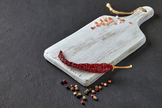 Red hot peppers peppercorns and coarse pink salt on a white wooden cutting board on a black concrete table Food display template