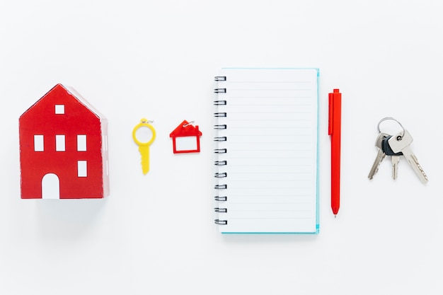 Red house model; plastic key; house shape keychain; spiral diary; pen and keys arranged in a row over white background