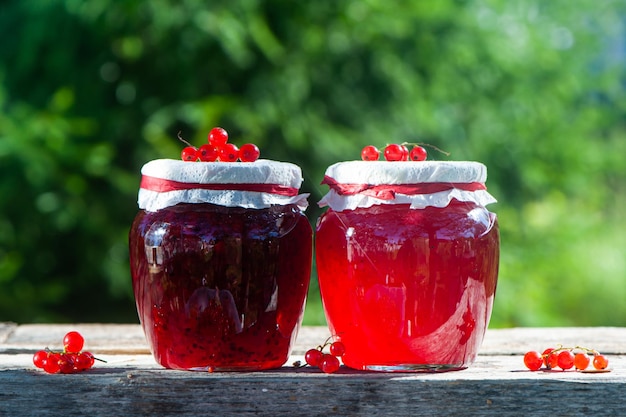 Red juicy berries of red currants and jars of berry jam on a\
wooden table on a green background of the garden harvest and\
cooking theme