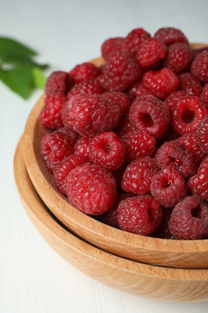 Red juicy raspberries in a wooden bowl on a white wooden background.