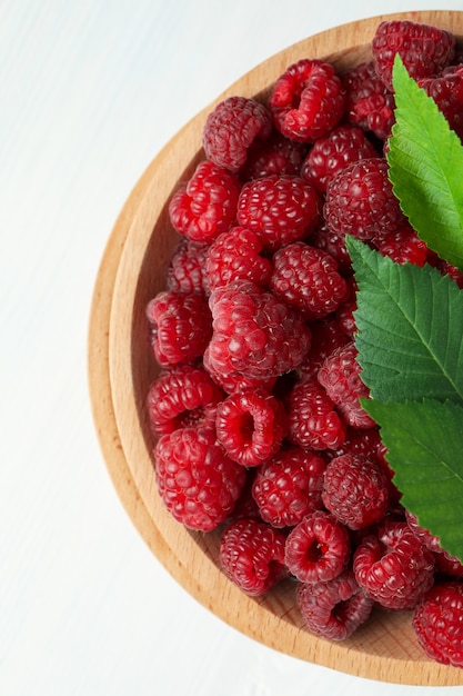 Red juicy raspberries in a wooden bowl on a white wooden background.