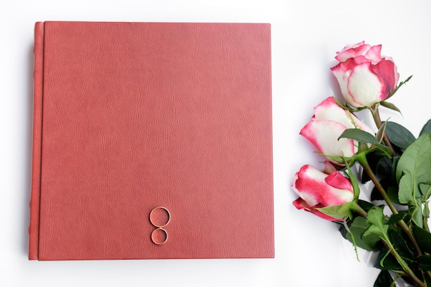 Red leather covered wedding book or album with two wedding rings and three roses lies on white background.