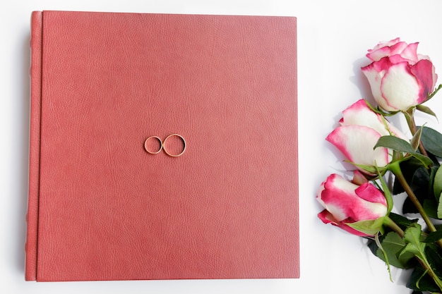 Red leather covered wedding book or album with two wedding rings and three roses lies on white background.