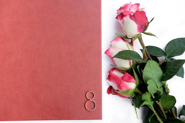 Red leather covered wedding book or album with two wedding rings and three roses lies on white background.