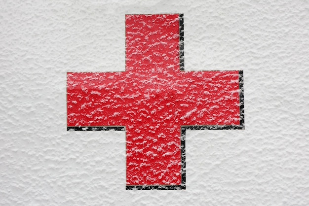 Photo red medical cross lightly covered with snow on white background outdoors.