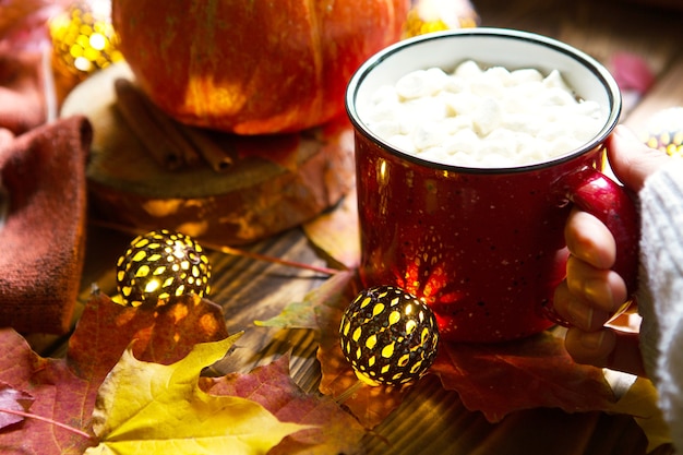 Foto tazza rossa di cioccolata calda con marshmallow sul tavolo con foglie di acero, ghirlande, bastoncini di cannella. atmosfera autunnale, caffè caldo, cacao, mani femminili in maglione bianco, intimità e comfort. copyspace