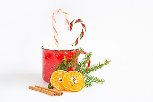 A red mug with marshmallows and candy cane in the shape of a heart is decorated with a natural fir branch or dried orange slices and cinnamon sticks. Christmas and New Year, the flavor of the holiday.