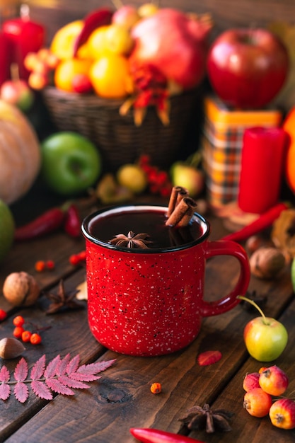 Red mug with mulled wine. Cinnamon sticks stick out of the cup and a star of star anise floats. Fruits and spices are all around on a wooden table