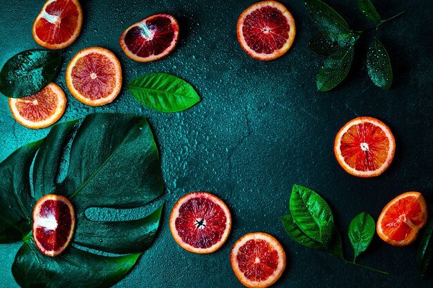 Красные апельсины, листья апельсинового дерева и лист монстера на темно-зеленом фоне под каплями воды.
