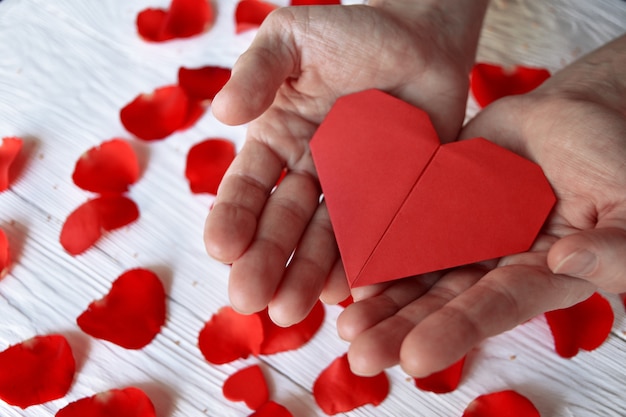 Red origami paper heart in male hands and rose petals. Valentine's Day concept