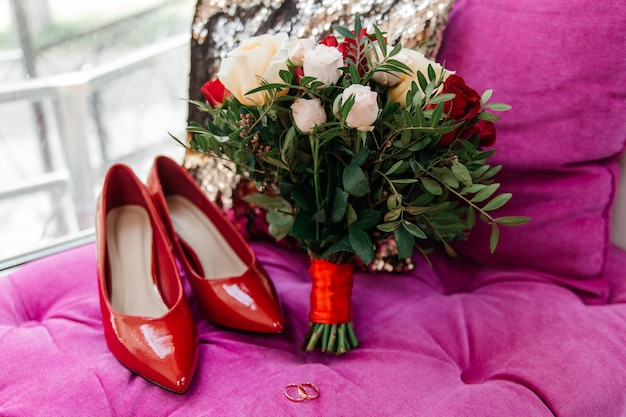 Photo red patent leather shoes of the bride near gold wedding rings and a bouquet of roses