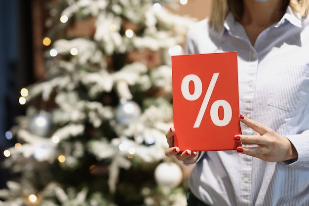 Red plaque with percent in female hands of seller against background of new year tree christmas