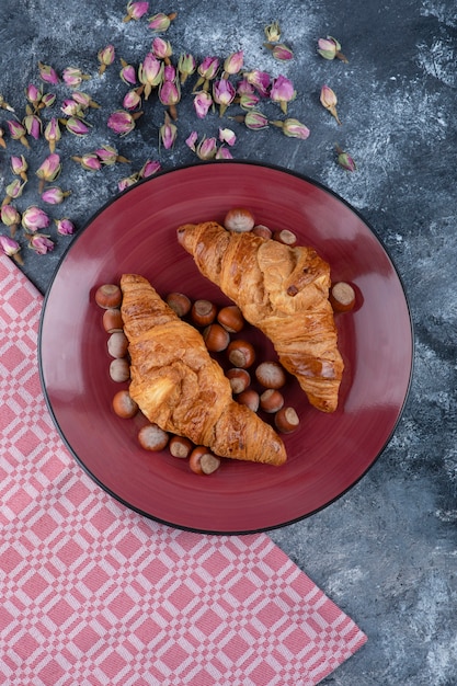 Red plate of sweet croissants with shelled hazelnuts on marble.