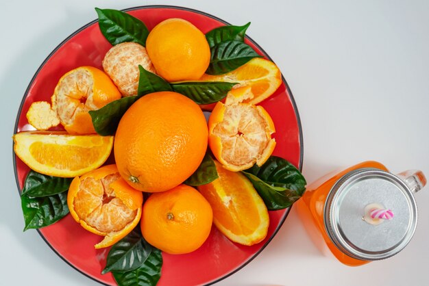 Red plate with oranges and tangerines green leaves bottle with juice on light background top view copy space