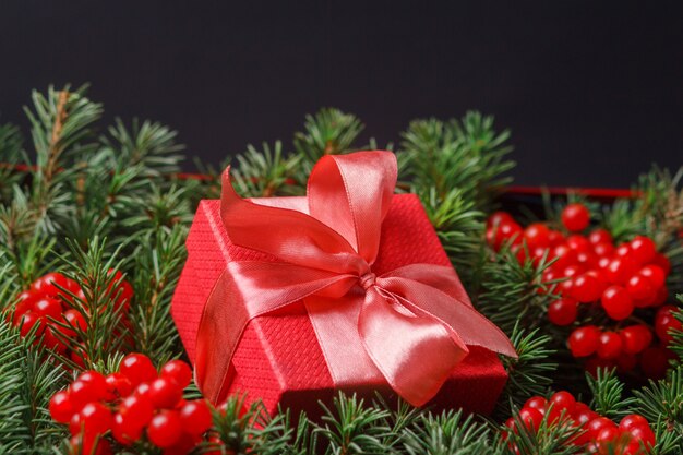 Red present gift box with satin pink bow, immersed in the needles of a Christmas tree decorated with red berries.