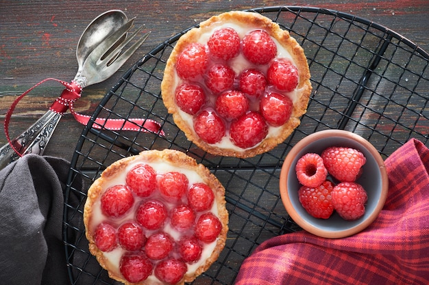 Red raspberry shortbread tarts with vanille custard and glazed fresh raspberries on cooling rack on dark brown wood