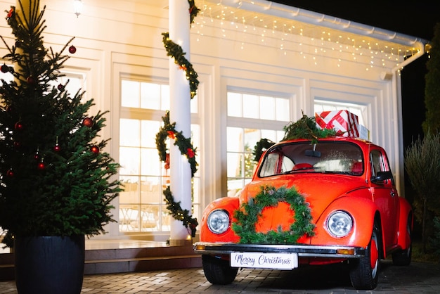 A red retro car decorated for Christmas with gifts on the roof stands near the house in the evening