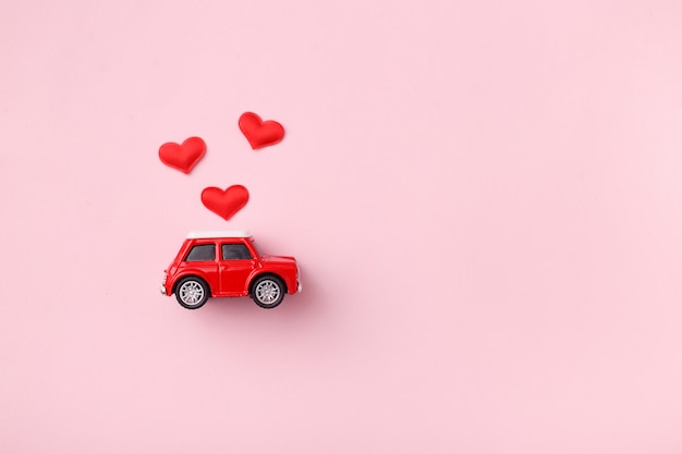 Red retro toy red car with red bow for Valentine's day on pink background with heart confetti. Top view, flat lay