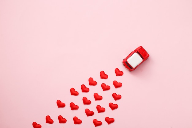 Red retro toy red car with red bow for valentine's day on pink background with heart confetti. top view, flat lay