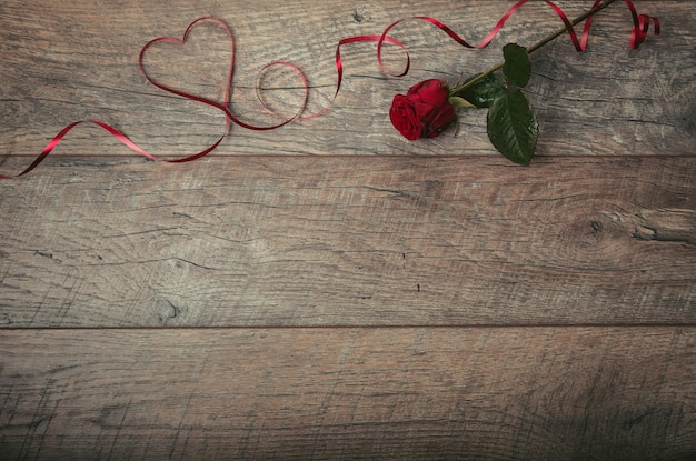 The red ribbon is lined in the shape heart on a dark wooden table. Valentines day background, rustic style. Valentines day table place setting with red rose, and ribbon. Flat lay, top view