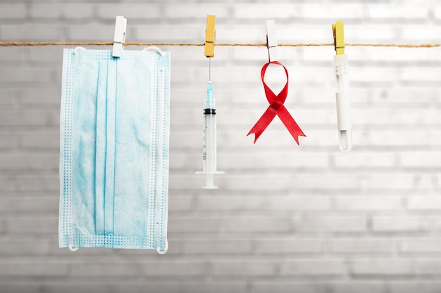Red ribbon, syringe, and face mask hanging on the rope with brick wall background. Hiv Aids ribbon awareness