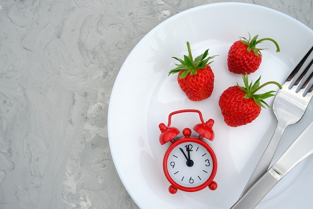 Red ripe strawberries berry on white plate, cutlery and red alarm clock on gray stone table, closeup